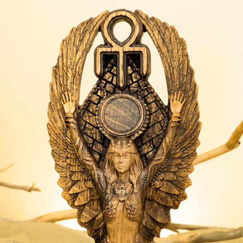 Może przedstawiać: Drewniana rzeźba skrzydlatego stworzenia z rozpostartymi ramionami, trzymającego symbol ankh nad piramidą. Postać otoczona jest okrągłą aureolą i ma szczeg&oacute;łową twarz.
