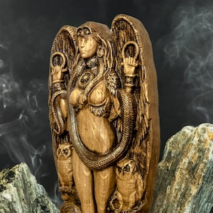 May include: A wooden statue of a winged goddess with a snake wrapped around her body. She is holding a ring in each hand and has owls on either side of her. The statue is painted in a golden brown color.