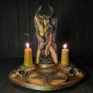 Könnte beinhalten: Ein dunkelbrauner Holzaltar mit einer zentralen geschnitzten Figur eines geflügelten Wesens, das von einer Schlange umschlungen ist und über einem Totenkopf thront. Die Basis enthält ein Pentagramm und zwei Kerzenhalter mit gelben Stumpenkerzen.