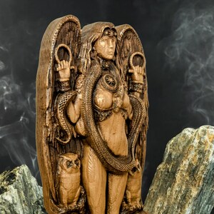 May include: A wooden sculpture of a female figure with wings, a snake wrapped around her body, and owls on either side. The figure is carved in a detailed style and has a warm brown color.