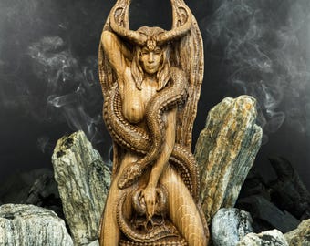 May include: A wooden sculpture of a woman with wings and horns, holding a snake in her arms. The sculpture is carved in a detailed style and has a dark brown finish.