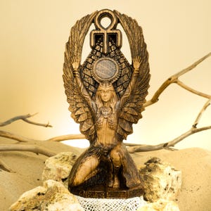 Może przedstawiać: Drewniana statua skrzydlatej bogini z rozpostartymi ramionami, trzymająca symbol ankh nad głową. Bogini klęczy i nosi koronę oraz biżuterię. Statua jest rzeźbiona z misternymi detalami i ma ciepły brązowy kolor.