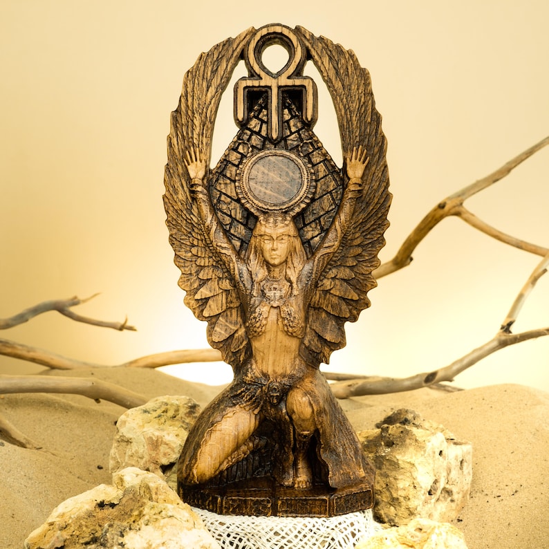Może przedstawiać: Drewniana statua skrzydlatej bogini z rozpostartymi ramionami, trzymająca symbol ankh nad głową. Bogini klęczy i nosi koronę oraz biżuterię. Statua jest rzeźbiona z misternymi detalami i ma ciepły brązowy kolor.