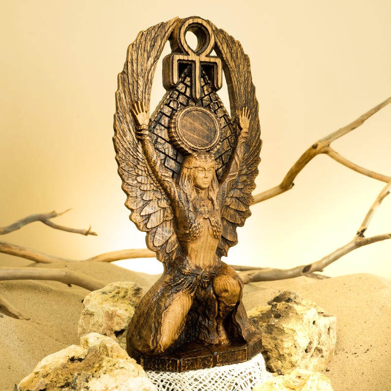 Może przedstawiać: Drewniana statua skrzydlatej bogini z rozpostartymi ramionami, trzymająca symbol ankh nad głową. Statua jest rzeźbiona z misternymi detalami i ma ciemnobrązowe wykończenie.