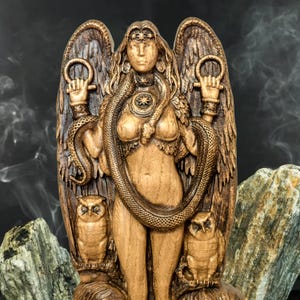May include: A wooden carving of a goddess figure with wings, a snake wrapped around her body, and owls on either side. The figure is holding a ring in each hand.