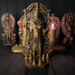 May include: A golden-brown, detailed sculpture of the Egyptian deity Anubis standing centrally, adorned with an ankh pendant, surrounded by intricate carvings of skulls and hieroglyphs, flanked by two lit pillar candles and other statues.