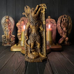 Peut inclure: Une statue brun doré de la divinité égyptienne Sobek, représentée avec une tête de crocodile et un corps humain, tenant un sceptre surmonté d'un ankh. La figurine repose sur un socle rectangulaire, entourée d'autres statues et deux bougies.