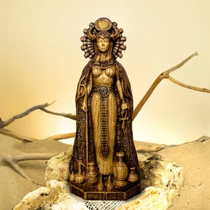 May include: A detailed, golden-colored statue of an Egyptian goddess. The figure wears a headdress, ornate cape, and holds a staff and an ankh. Several decorative vases are at the base of the statue, which is set on a stone surface.