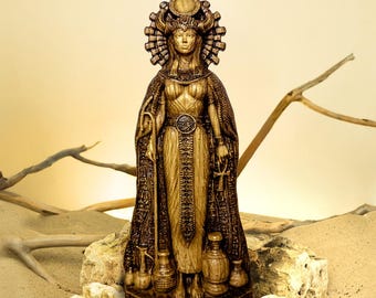 Puede incluir: Estatua dorada detallada de una diosa egipcia. La figura lleva una toca, una capa adornada y sostiene un báculo y un anj. Varios jarrones decorativos están en la base de la estatua, colocada sobre una superficie de piedra.