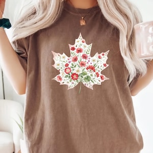 Op de afbeelding: Bruin t-shirt met een bloemig esdoornbladontwerp. Het blad is gevuld met rode, witte en groene bloemen. Het shirt heeft korte mouwen en een ronde hals.