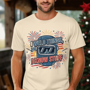 May include: A cream-coloured t-shirt with a blue welding mask graphic, sunglasses, fireworks, and American flag details. The text on the shirt reads "I WELD THINGS, I KNOW STUFF".
