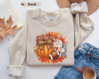 May include: A sand-colored crewneck sweatshirt featuring a graphic print of an orange pumpkin with a leopard print bow, a plaid blanket, a football, a plastic cup with a straw, a lantern, and autumn leaves in shades of red, orange, and yellow.