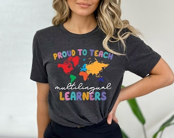 May include: A dark heather gray short-sleeve t-shirt featuring a colorful world map graphic. The text reads PROUD TO TEACH in a multi-colored font above the map, and multilingual LEARNERS in a script and block font below the map graphic.