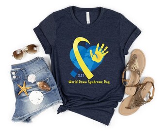 May include: A dark navy blue t-shirt featuring a graphic of a blue heart, a yellow ribbon, and a yellow handprint. The text "3.21" and "World Down Syndrome Day" are printed in yellow below the heart. The shirt is displayed with denim shorts, sunglasses, and sandals.