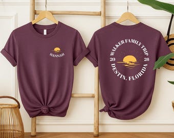 May include: A dark plum t-shirt featuring a small sunset graphic and the name HANNAH on the front left chest. The back displays a large circular graphic with a sunset, the text WALKER FAMILY TRIP, DESTIN, FLORIDA, and the year 2026.