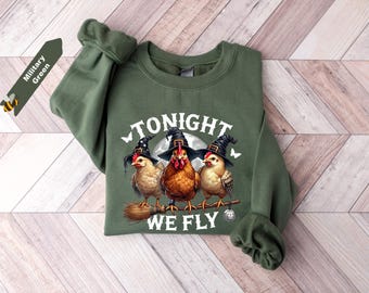 May include: A military green crewneck sweatshirt featuring a graphic of three chickens wearing black witch hats. The chickens are perched on a broomstick with a disco ball hanging from it. The text TONIGHT WE FLY is printed in white.