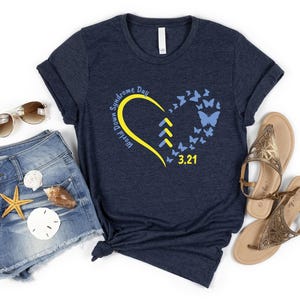 May include: A dark navy blue short-sleeve t-shirt featuring a yellow heart outline, three yellow chevron arrows, and blue butterflies. The text reads World Down Syndrome Day and 3.21. The shirt is displayed with denim shorts, sunglasses, and sandals.