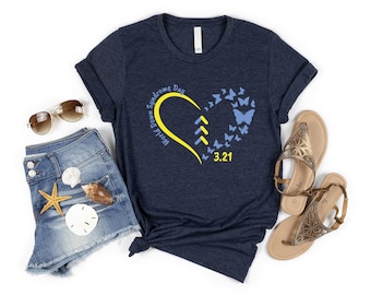 May include: A dark navy blue short-sleeve t-shirt featuring a yellow heart outline, three yellow chevron arrows, and blue butterflies. The text reads World Down Syndrome Day and 3.21. The shirt is displayed with denim shorts, sunglasses, and sandals.