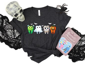 May include: A dark heather gray t-shirt featuring a graphic of four cartoon teeth dressed as Halloween characters: a green Frankenstein, a white ghost, a white mummy, and an orange jack-o'-lantern, all surrounded by small white bats.