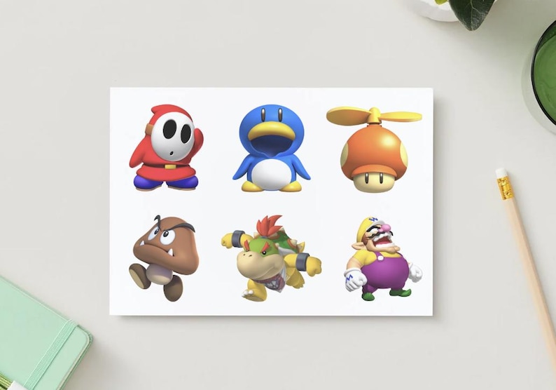 May include: A white card featuring six colorful cartoon characters from the Super Mario video game series. The characters include Shy Guy, a blue penguin, a mushroom with a propeller, a Goomba, a Koopa, and Wario.