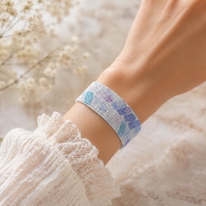 Könnte beinhalten: Ein Perlenarmband mit weißer Basis und einem Muster aus blauen und lila Formen. Das Armband wird am Handgelenk getragen, und das Design ist auf der Haut sichtbar. Das Armband ist etwa 3,8 cm breit.