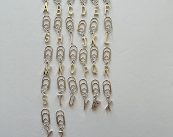 May include: A collection of silver-toned paper clips, each adorned with a different letter of the alphabet in a gold color. The letters are suspended from the paper clips, creating a decorative and personalized stationery item. The background is a plain white surface.