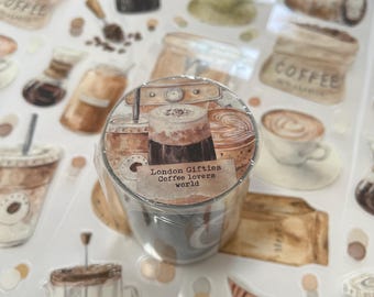 Può includere: Un rotolo di nastro adesivo decorativo con illustrazioni a tema caffè, tra cui tazze, chicchi e sacchi. Il nastro ha il testo "London Gifties Coffee lovers world" stampato sopra. Lo sfondo è pieno di adesivi simili a tema caffè.