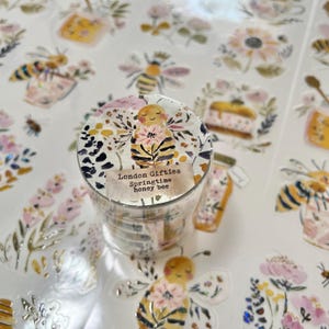 May include: A roll of decorative washi tape featuring a springtime honey bee design. The tape has a floral and bee pattern with the text "London Gifties Springtime honey bee". Surrounding the tape are bee and floral stickers.