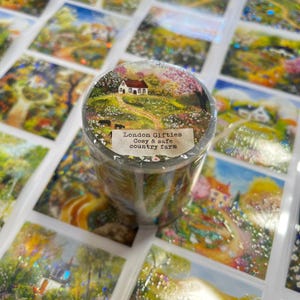 May include: A roll of decorative tape featuring a countryside landscape with a white house, a winding path, and cows. The tape has the text "London Gifties Cosy & safe country farm" on a label. The background shows other landscape images.