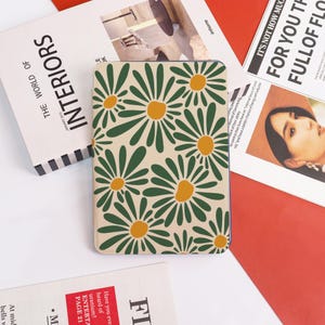 May include: A beige tablet case featuring a repeating pattern of green daisies with yellow centers. The case is rectangular with rounded corners. The words "INTERIORS" and "THE WORLD OF" are visible on a magazine in the background.