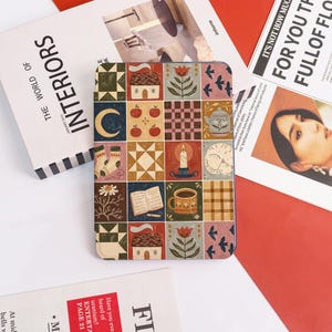 May include: A patterned tablet case featuring a patchwork design with various illustrations. The squares depict a crescent moon, apples, a house, a teacup, and other cozy motifs. The case is surrounded by magazines and a book.
