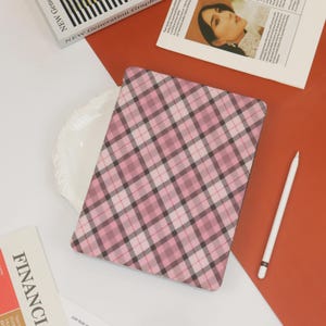 May include: A pink and black plaid tablet case with a white stylus. The case is rectangular and features a diagonal plaid pattern in shades of pink and black. The stylus is white with a black tip. The background includes a white plate and a red surface.