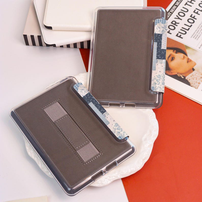 May include: Clear protective case for an e-reader with a brown interior and a patterned fabric strap. The case has a built-in stand for hands-free use. The design includes a clear plastic frame and a decorative fabric accent.