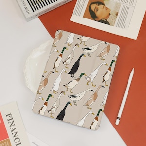 May include: A beige tablet case featuring a repeating pattern of stylized ducks in various colors, including white, brown, and black. The case is rectangular and lies on a surface with other items, including a white stylus and a magazine.