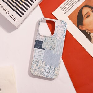 May include: A phone case with a patchwork design in shades of blue and white. The case features various floral and geometric patterns, creating a textured appearance. The case is on a white surface with a red background and a book.