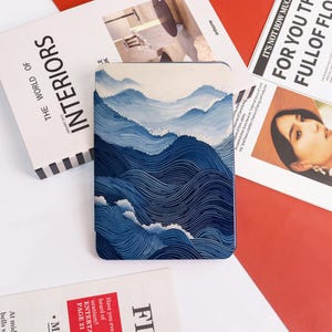 May include: A rectangular case with a blue and white wave design. The case features a stylized depiction of ocean waves and mountains. The case is surrounded by magazines and newspapers.