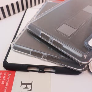 May include: Two clear phone cases with a gray interior and a built-in stand are stacked on top of a black and white surface. The cases have cutouts for buttons and ports. The bottom case has a black border.