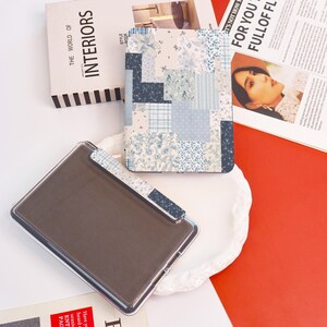 May include: A clear plastic case with a dark brown interior and a patterned clip. A book titled "The World of Interiors" and a patterned notebook with blue floral and patchwork designs are also visible.