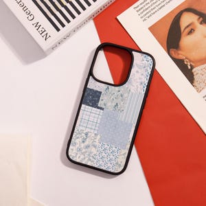 May include: A black phone case featuring a patchwork design in various shades of blue and white. The case has a black frame and a floral pattern. The phone case is on a white surface, next to a book and a magazine.