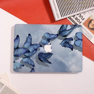 May include: A laptop case featuring a design of blue butterflies on a light blue background. The case has a white Apple logo in the center. The butterflies vary in size and shade, creating a whimsical aesthetic.