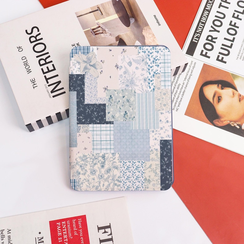 May include: A patchwork-style tablet case featuring a variety of blue and white floral and geometric patterns. The case is surrounded by magazines and publications, including one titled "INTERIORS". The case has a dark blue border.