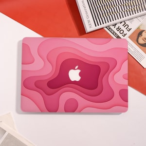 Könnte beinhalten: Eine rosa Laptop-Hülle mit einem geschichteten Design in Rosatönen, mit einem weißen Apple-Logo. Die Hülle hat eine glatte Oberfläche und eine moderne Ästhetik. Die Hülle liegt auf einer weißen Oberfläche mit rotem Hintergrund.