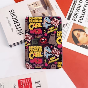 May include: A rectangular e-reader case featuring a black background with a repeating pattern of Dungeon Crawler Carl graphics, including blue dinosaurs, gray cats, red lips, and text that reads New Achievement and God Damnit Donut.