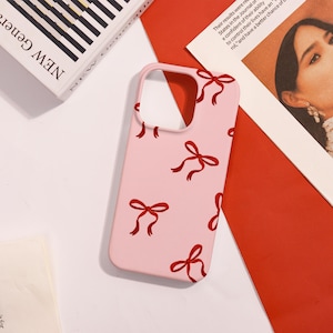 May include: A pink phone case featuring a pattern of red bows. The case has cutouts for the camera and buttons. The phone case is on a white and red surface.