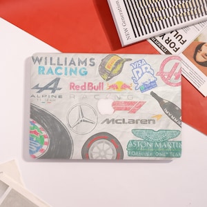 May include: A light gray laptop skin featuring Formula 1 racing team logos and text, including "Williams Racing," "Red Bull," and "McLaren." The design incorporates a tire graphic and the Apple logo.