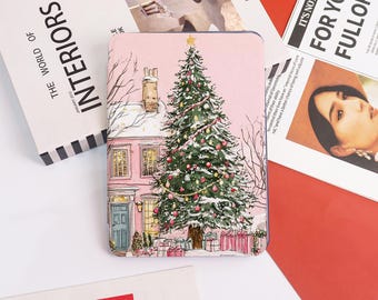 May include: A pink compact mirror with a Christmas scene. The illustration features a decorated Christmas tree in front of a pink house with a blue door. Presents are at the base of the tree. The mirror is surrounded by magazines.