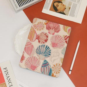 May include: A white iPad case with a colorful seashell pattern. The case features a variety of seashells in shades of pink, blue, red, and yellow.