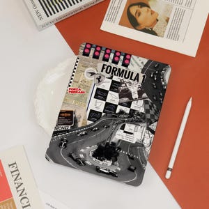 May include: A notebook with a Formula 1 theme, featuring a black and white race track design. The notebook has the text "FORMULA 1" and "FORZA FERRARI". A white stylus and other printed materials are also visible.
