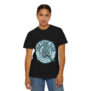 May include: A black short-sleeve t-shirt featuring a circular graphic with a light blue border. The design includes a magnifying glass over a fingerprint, the text COLD CASE CLUB, and the phrase MEMBER SINCE FOREVER in a distressed font.