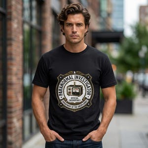 May include: A black short-sleeved t-shirt featuring a circular graphic on the chest. The design includes the text "PARANORMAL INVESTIGATOR" and "OFFICIAL CREDENTIALS" surrounding an illustration of a vintage radio device with a gauge.